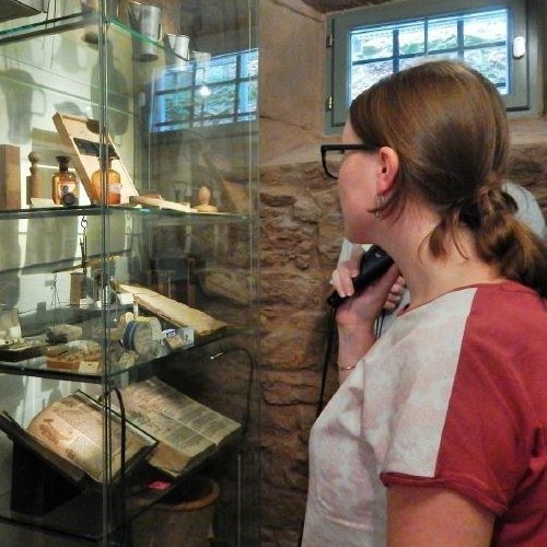 Eine junge Frau hält einen Audioguide in der Hand. Währendessen schaut sie auf eine Vitrine mit Ausstellungsstücken.