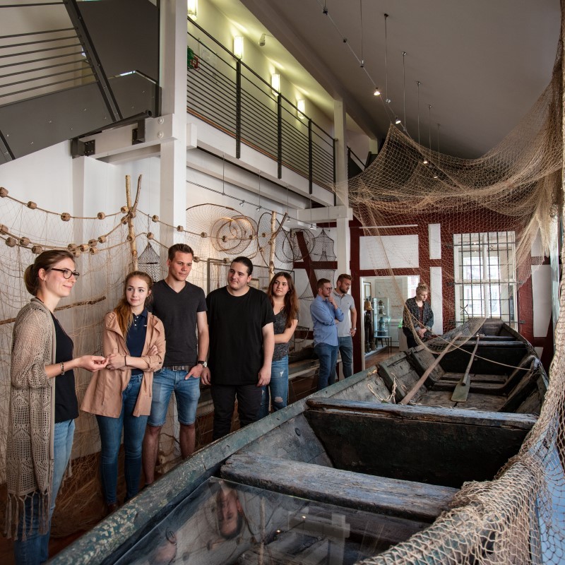 Eine Gruppe von Erwachsenen besucht eine Führung durch die Ausstellungsräume des Museum Stadt Miltenberg. Sie befinden sich in der Fischereiabteilung mit einem alten Fischerboot.